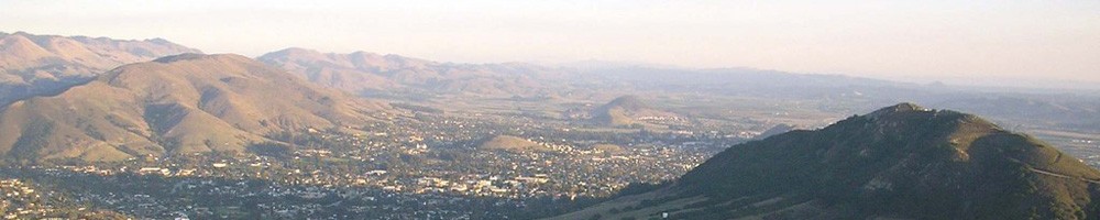 Panoramic view of San Luis Obispo from the surrounding hills at golden hour, with the city nestled in a valley between green and golden mountains